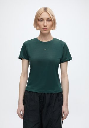 Kobieta o krótkich blond włosach, ubrana w dopasowany ciemnozielony t-shirt i luźne czarne spodnie, stoi na tle gładkiego jasnego tła.