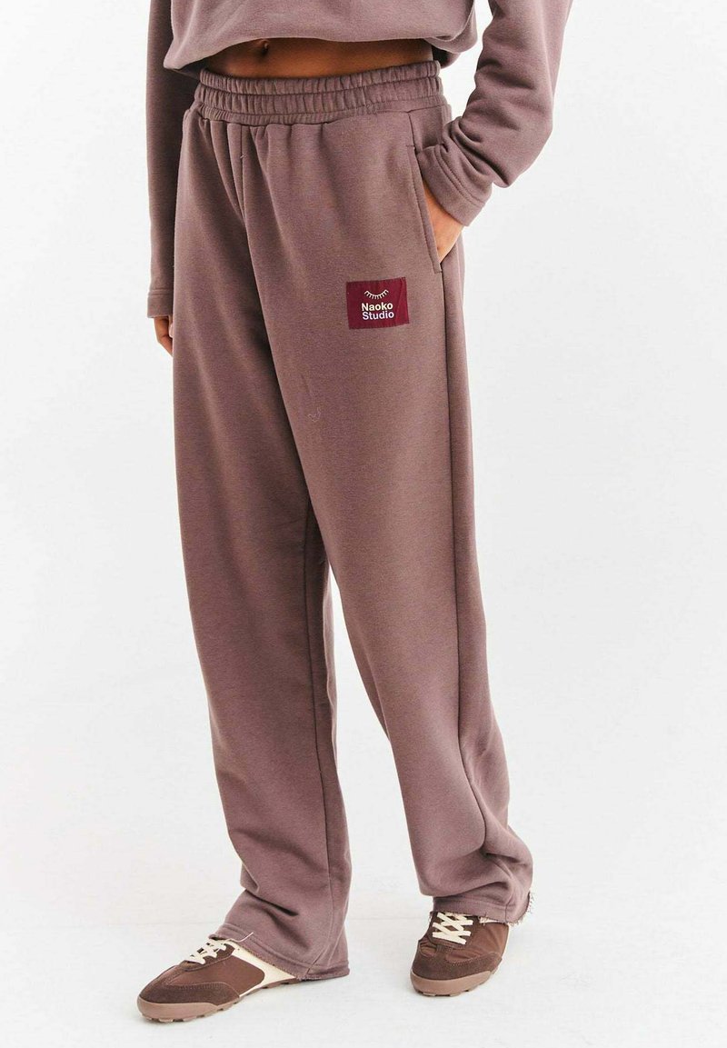 Brązowe spodnie dresowe wykonane z miękkiego materiału, z elastycznym pasem, bocznymi kieszeniami oraz małą bordową metką z logo "Naoko Studio".
