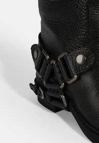 Zwarte leren enkelboot met textuurmateriaal, metalen gespen en een lage gestapelde hak. Kenmerken zijn onder andere een gewikkeld bandontwerp en zilveren hardware.