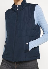 Navy blue puffer vest with a high collar, zip front, and side pockets. Features quilted texture and metallic snap accents.