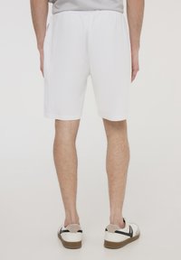 Pantaloni shorts bianchi in cotone con una cintura elastica, lunghi fino al ginocchio, che presentano una trama liscia. Indossati con scarpe da ginnastica casual bianche e beige.