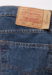 Jeans i denim med en brun läderetikett med r öd tryck. Etiketten visar "Levi Strauss & Co." och detaljer inkluderande "Lot 501, W 32 L 32."