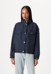 Giacca blu accorciata realizzata in materiale leggero, con due tasche frontali, chiusura a scatto, colletto alto e polsini regolabili.