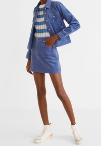 Completo giacca e gonna in corduroy blu con una maglietta a righe. Le caratteristiche includono dettagli con bottoni e sneakers bianche alte. Texture liscia.