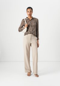Leopardmønstret skjorte med knapper, beige vide bukser og lysebrune sandaler. Modellen holder en hvid skuldertaske foran en ensfarvet baggrund.