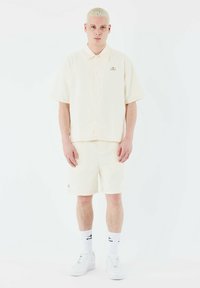 Conjunto de camiseta de manga corta con collar y pantalones cortos en un suave color crema, fabricado con un tejido texturizado con sutiles líneas verticales; presenta un pequeño logotipo en la camiseta.