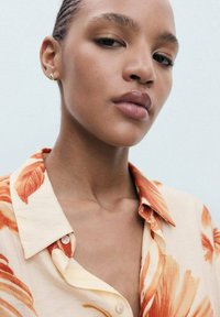 Collared shirt featuring orange floral print on a light background, made from a textured fabric. Model displayed with minimal accessories.