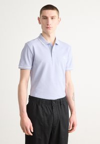 Hellblaues Poloshirt mit Kragen, weißen Besätzen und kurzen Ärmeln. Mit einem Logo auf der Brust. Kombiniert mit schwarzen Hose mit elastischem Bund.