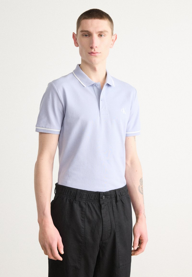 Hellblaues Poloshirt mit Kragen, weißen Besätzen und kurzen Ärmeln. Mit einem Logo auf der Brust. Kombiniert mit schwarzen Hose mit elastischem Bund.