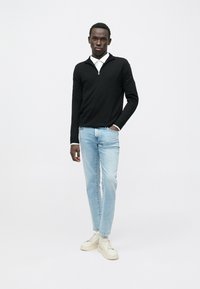 Schwarzer Pullover mit langen Ärmeln und halbem Reißverschluss am Hals, getragen über einem weißen Hemdkragen, kombiniert mit hellblauen Jeans und beigen Turnschuhen.