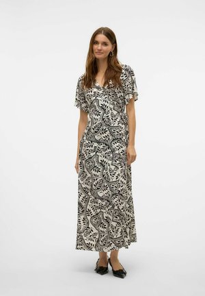 Femme aux longs cheveux bruns portant une robe portefeuille à motifs noir et blanc et des chaussures noires à bout pointu, se tenant devant un fond blanc uni.