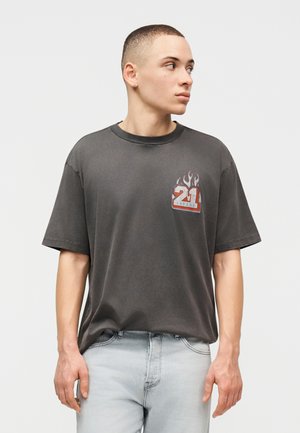 Ung mand i grå grafisk t-shirt med "21 Savage" og flammedesign, kigger mod højre, iført lyseblå jeans.