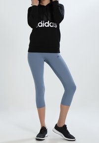 Sudadera negra con capucha y logo blanco de "adidas", combinada con leggings recortados azul claro y zapatillas deportivas negras.