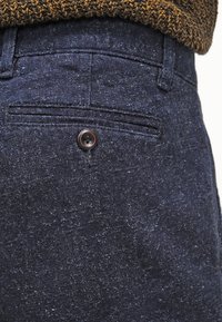 Dark blue textured fabric pant featuring a rear pocket with a single brown button. Smooth finish with a subtle pattern visible.