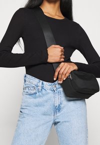 Schwarze Stoff-Crossbody-Tasche mit Reißverschluss, rechteckiger Form und glatter Textur; kombiniert mit hellblauen hochtaillierten Jeans und einem schwarzen Oberteil.