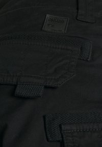 Mustapohjaiset cargo-housut, joissa on useita taskuja ja teksturoitu kangas, johon on merkitty "Alpha Industries". Ompeleet parantavat kestävyttä.