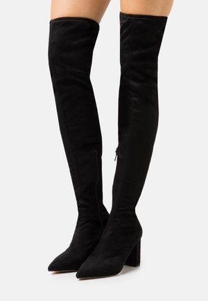 Svart mockastövel som går upp till knäet med spetsig tå och klumpig klack; har en sidodragkedja för stängning och en slät yta.