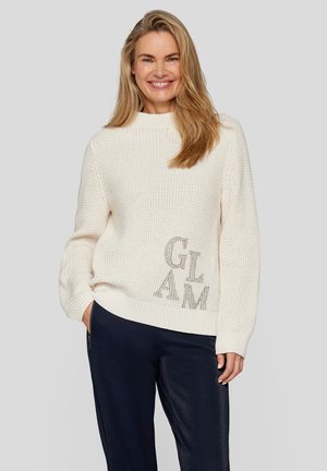 Cream knitted sweater with a high neckline and ribbed cuffs. Features "GLAM" in metallic lettering on the front. Paired with dark blue pants.