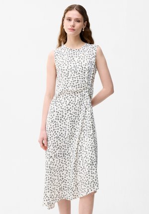 Kobieta w białej sukience midi bez rękawów z czarnym nieregularnym wzorem kół, stoi z jedną ręką za plecami.