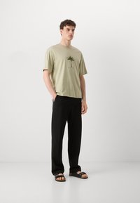 Lysegrøn bomulds t-shirt med et palmetræsgrafik, parret med sorte bukser og sorte sandaler. Modellen står med hænderne i lommerne.