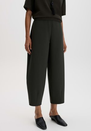 Personne portant un pantalon large court de couleur foncée, des mules noires à enfiler et un haut sombre à manches courtes avec un collier à pompons.