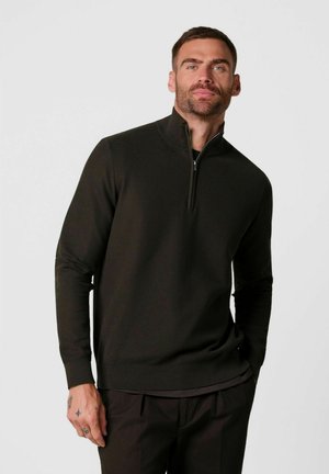 Uomo che indossa un pullover nero a maniche lunghe con una cerniera a un quarto, in piedi su uno sfondo chiaro e semplice con le mani rilassate.