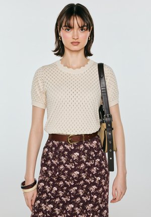 Młoda kobieta z krótkimi ciemnymi włosami nosi kremowy dzianinowy top z krótkim rękawem, kwiecistą spódnicę, brązowy pasek, nosi beżową i czarną torbę na ramię oraz nosi bransoletki.