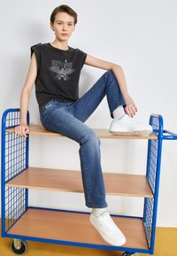 Canotta nera con grafica di un'aquila, jeans blu e sneakers bianche con suole testurizzate, seduto su uno scaffale di metallo blu.