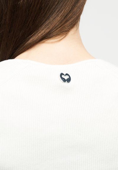 Μακριά καστανά μαλλιά πάνω από ένα λευκό πλεκτό πουκάμισο με ριμπ, που εμφανίζει ένα μικρό μεταλλικό κούμπωμα σε σχήμα καρδιάς στο πίσω μέρος του λαιμού.