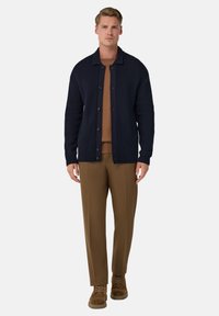 Marinblå ribbad kofta med knappar, bärs över en brun tröja. Matchas med bruna byxor och bruna skor, stående mot en vit bakgrund.