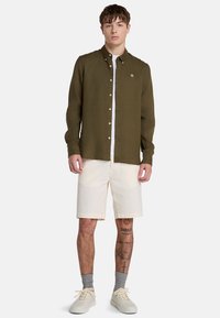 Camisa verde oliva, de botones y de tela texturizada. Lleva puesta una camiseta blanca y unos pantalones cortos crema. Unas zapatillas de deporte de color claro completan el conjunto.