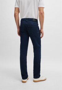 Jeans din denim albastru închis, cu o croială clasică, având două buzunare posterioare și un petic cu logo negru pe talie, asortați cu adidași albi.