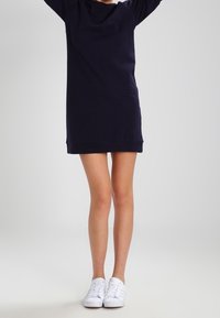 Navy-blaues Sweatkleid mit langen Ärmeln, geripptem Saum und weicher Textur, kombiniert mit weißen Sneakers, vor einem neutralen Hintergrund präsentiert.