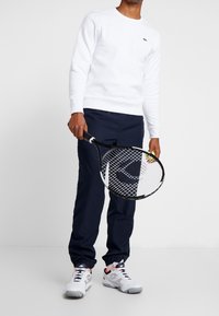 Vit sweatshirt med rund hals, marinblå byxor och vita tennisskor. Håller en tennisracket med en neongul boll.