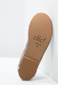 Zool van een beige platte schoen met "clic!" logo, maat 31, gemaakt in Spanje, leunend tegen een witte hoekmuur.