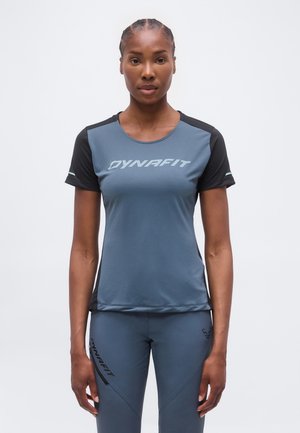 Γυναίκα φορώντας μπλε και μαύρο αθλητικό μπλουζάκι Dynafit και αντίστοιχο μπλε παντελόνι, στέκεται μπροστά σε απλό φωτεινό φόντο.