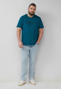 Turquoise katoenen t-shirt met een ronde hals, met een donker gedrukt logo. Gepaard met lichtblauwe jeans en witte schoenen met een beige zool.