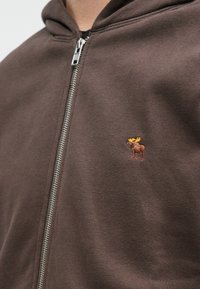 Pruun tõmblukuga kapuutsiga t-särk, mis on valmistatud pehmest kangast, mille vasakul rindkereosal on väike tikitud põdra logo. Hõbedane tõmblukk.