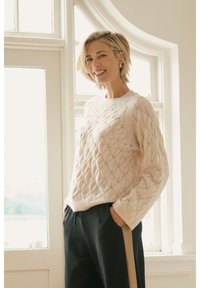 Pull beige clair avec un motif en losange texturé, col rond, manches longues, associé à un pantalon noir avec une bande latérale.