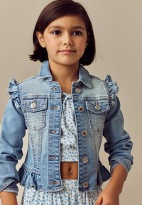 Junges Mädchen mit kurzen braunen Haaren, das ein blaues geblümtes Crop-Top-Kleid und eine hellblaue Jeansjacke mit gerafften Schultern trägt.