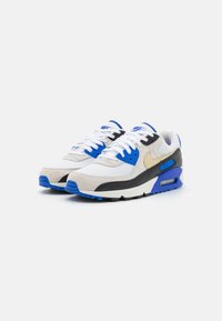 Um par de ténis Nike Air Max em branco, azul, bege e preto, com atacadores, detalhes em malha e camurça, sobre um fundo branco.
