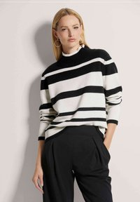 Gestreiftes Sweatshirt in Schwarz und Weiß mit hohem Kragen und gerippten Bündchen, aus einem weichen Strickmaterial. Kombiniert mit schwarzen Hosen.