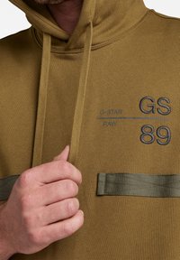 Sudadera verde oliva con capucha ajustable, que cuenta con un bolsillo frontal y un logo bordado de "G-STAR" en gris oscuro con el número "89".