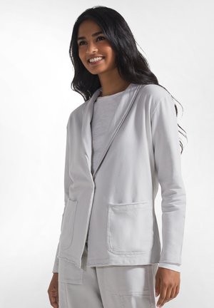 Kvinde med langt sort hår, smilende, iført lys grå afslappet blazer med lommer og matchende bukser mod en ensartet hvid baggrund.