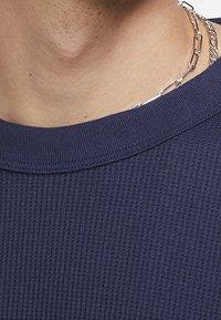 Námořnická pruhovaná košile s širokým kulatým výstřihem a hladkou texturou. Obsahuje detail stříbrného řetízku na pokožce.