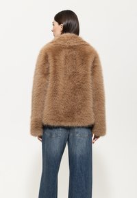 Braune Kunstfelljacke mit einer weichen, strukturierten Oberfläche, großem Kragen und weiten Ärmeln, kombiniert mit hellblauen, weit geschnittenen Jeans.