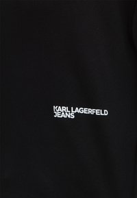 Μαύρο ύφασμα με λευκό κείμενο που γράφει "KARL LAGERFELD JEANS", τοποθετημένο στο κέντρο του κάτω μέρους της ορατής περιοχής.