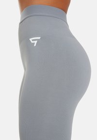 Grijze high-waisted leggings van rekbaar materiaal, met een wit logo aan de linkerkant en een geribbelde textuur langs de tailleband.