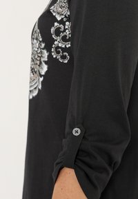 Sort langærmet bluse med et dekorativt blomstermønster i grå og creme, med en opsmøgning, der er sikret med en knapdetalje.