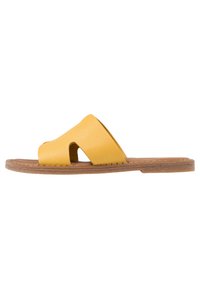 Gul lädersandal med platta sulor och två utskurna sektioner, med brun platt sula och stickningar längs kanterna för extra detalj och stabilitet.